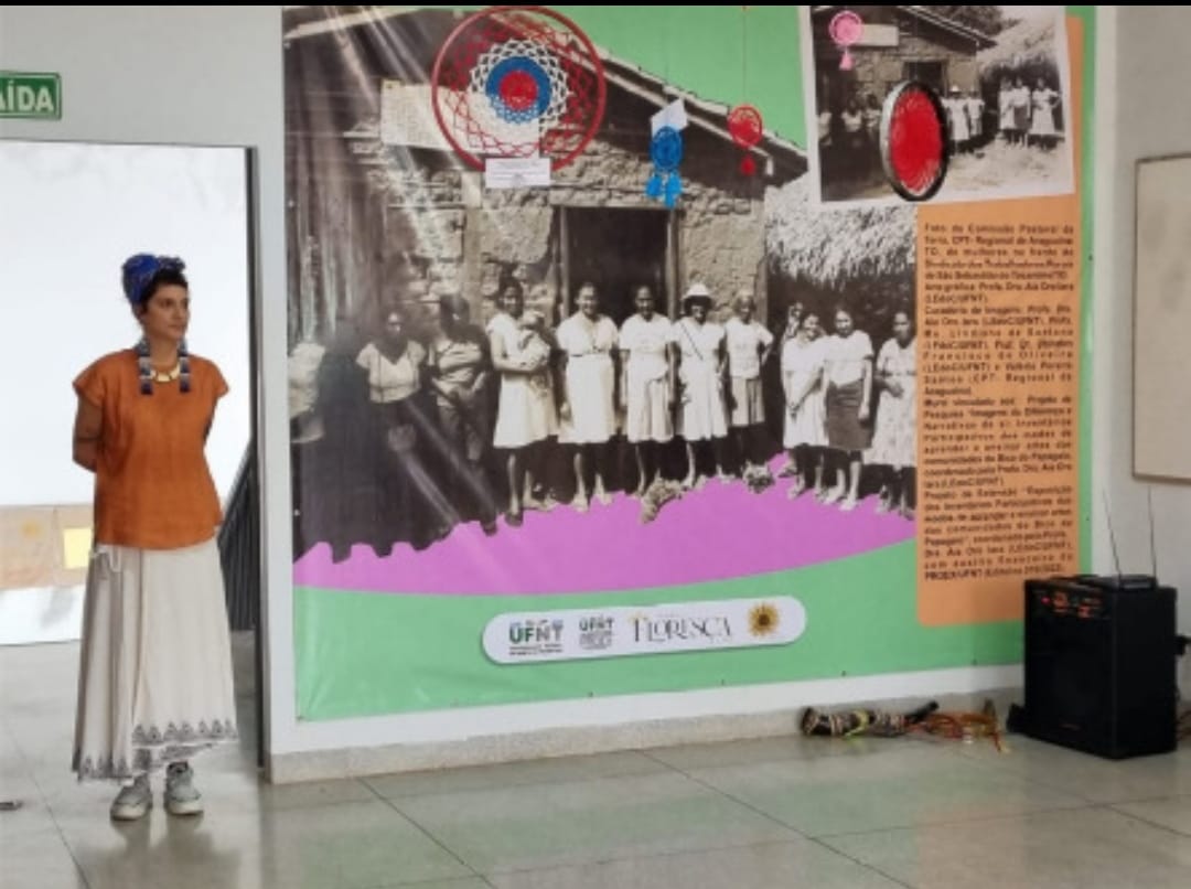 Figura 4: Professora Aia Oro Iara ao lado de um dos murais com fotos históricas cedidas pela CPT/Regional de Araguaína/ TO onde vemos um grupo de mulheres na frente do Sindicato dos Trabalhadores Rurais, de São Sebastião do Tocantins/ TO. Foto: Nataniel  Araújo, vice-reitor da UFNT, 2022.